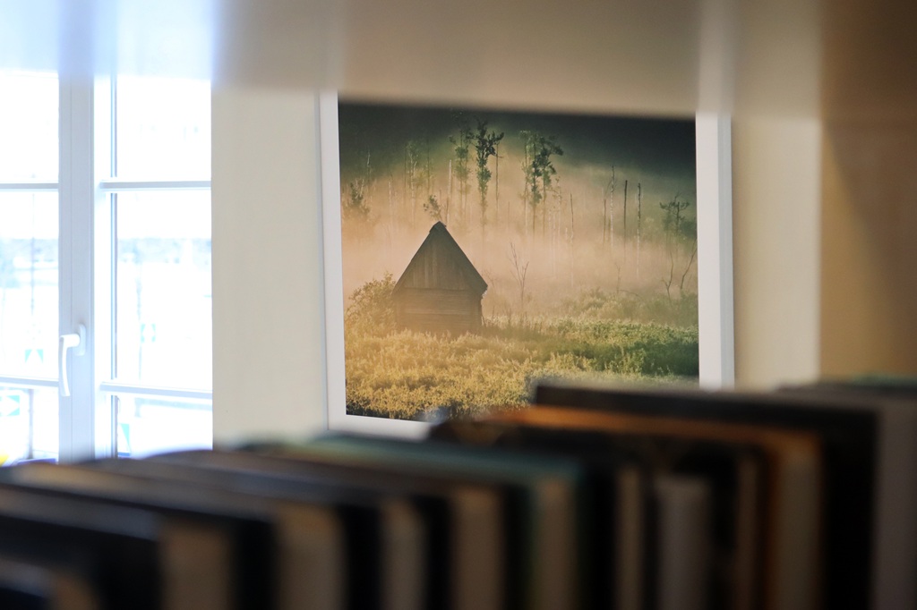 Caur grāmatu plauktu redzams ierāmēts miglainas ainavas attēls ar nelielu būdu un kokiem, radot mieru. No tuvējā loga iespīd saules gaisma.