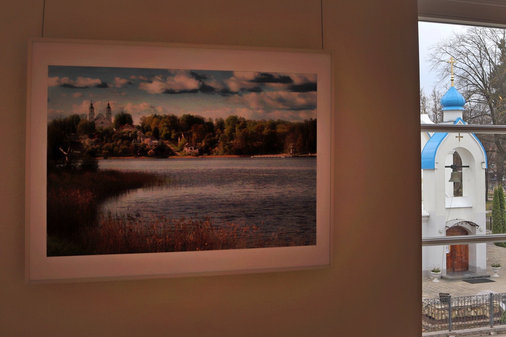 Pie sienas karājas ierāmēta ainavas glezna, kurā attēlots ezers ar ciematu un mākoņainām debesīm. Caur logu redzama balta baznīca ar zilu kupolu.