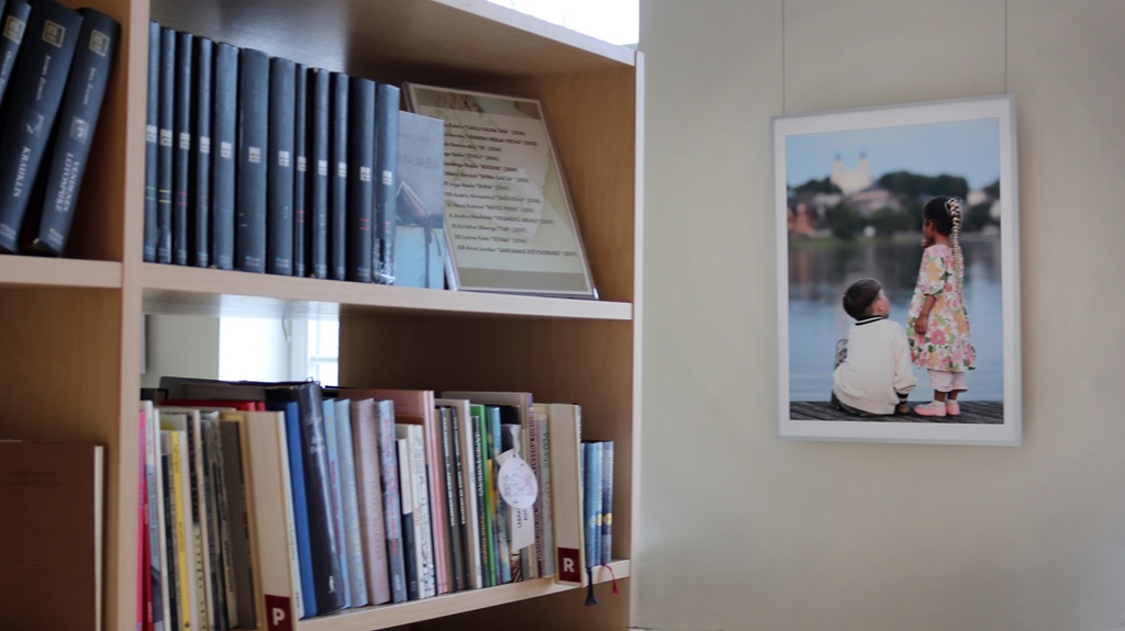 Pie sienas stāv koka grāmatu plaukts, kas pilns ar dažādām grāmatām, tostarp dažām zilos vākos. Pie sienas karājas divu bērnu fotogrāfija pie ezera.