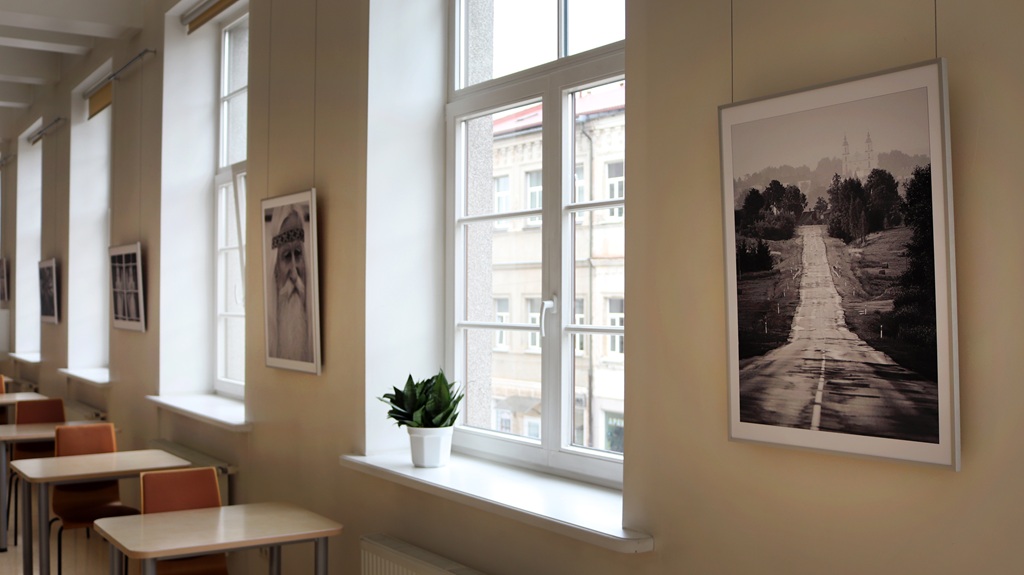 Gaiša lasītava ar lieliem logiem un ierāmētām melnbaltām fotogrāfijām uz smilškrāsas sienām, kurās attēlots garš ceļš. Podā audzēts augs uz palodzes.