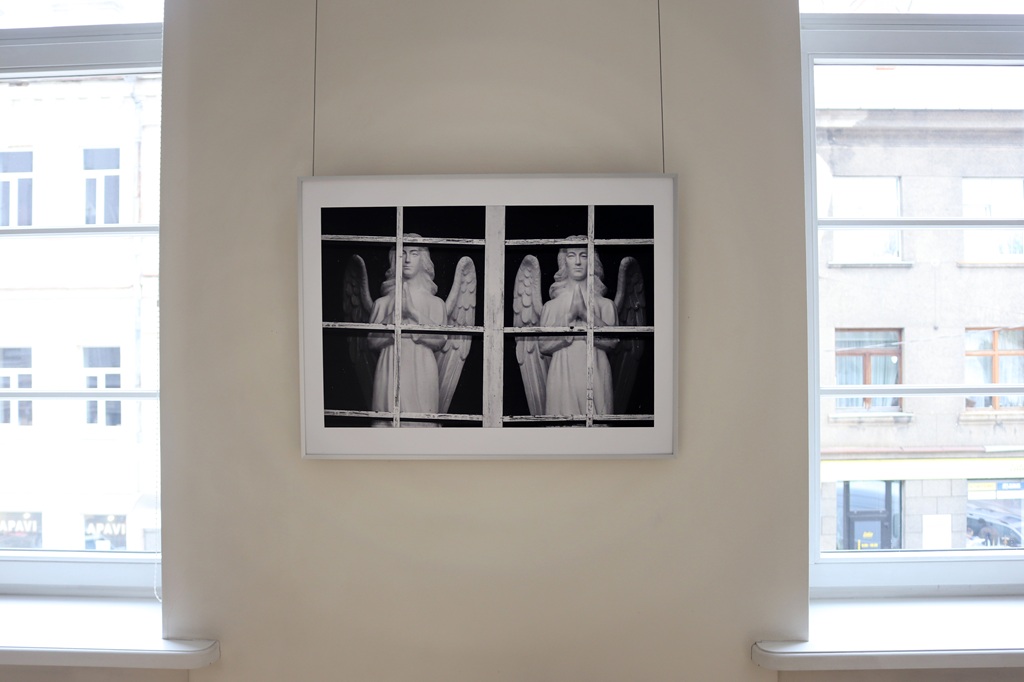 Ierāmēta melnbalta fotogrāfija ar divām eņģeļu statujām aiz logu rūtīm, kas piestiprinātas pie sienas starp diviem logiem. Aina rada mierīgu, pārdomu pilnu noskaņu.