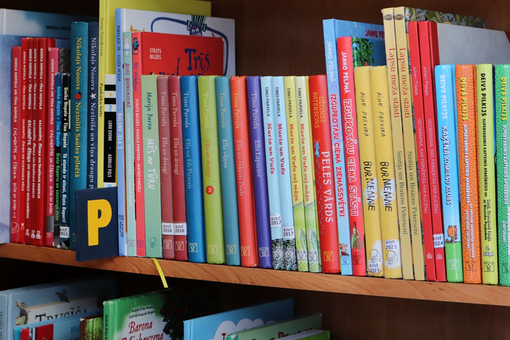 Grāmatu plaukts, kas pilns ar dažāda izmēra krāsainām bērnu grāmatām. Spilgti sarkani, zili, dzelteni un zaļi toņi rada dzīvīgu, aicinošu atmosfēru.