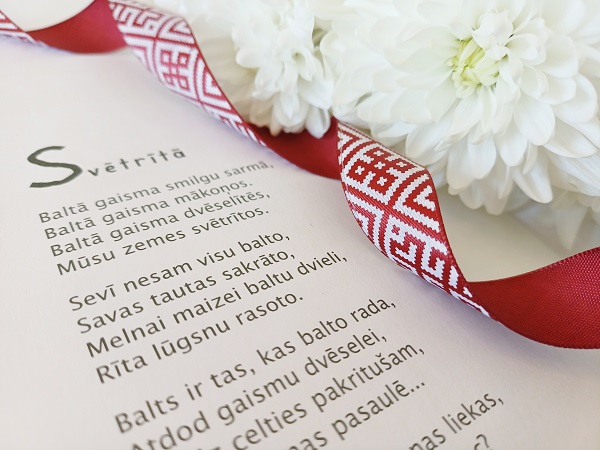 Dzejoļa lapas tuvplāns ar tekstu, kas dekorēts ar baltiem ziediem un sarkanu lentīti, kurā attēloti tradicionālie latviešu raksti, paužot eleganci un kultūras nozīmi.