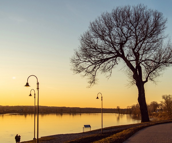 Mierīga upes krastmala saulrietā ar lielu, bezlapu koku un ielu laternām gar līkumotu celiņu. Garām paiet pāris, radot mierīgu un pārdomu pilnu noskaņu.
