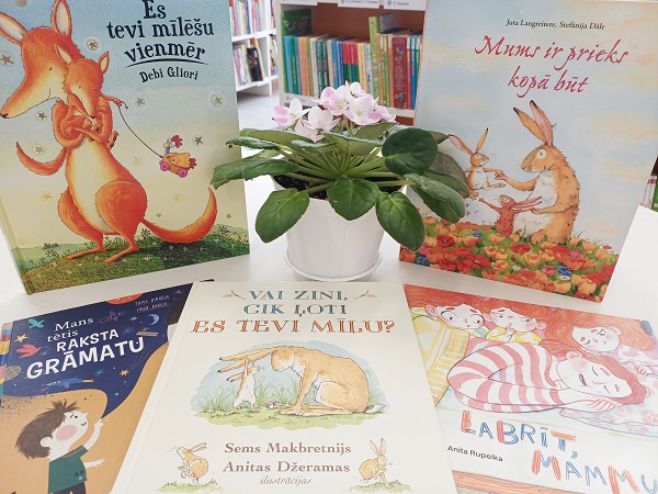 Piecas bērnu grāmatas puslokā izvietotas ap podaugu ar purpursarkaniem ziediem. Fonā redzams grāmatu plaukts bibliotēkā. Aina ir krāsaina un aicinoša.