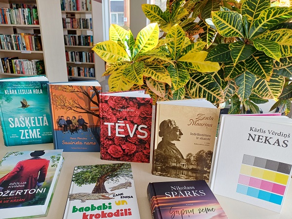 Grāmatas, kas izliktas uz galda bibliotēkā ar lapu augu fonā. Spilgti vāki un mierīga atmosfēra rada atklāšanas sajūtu.