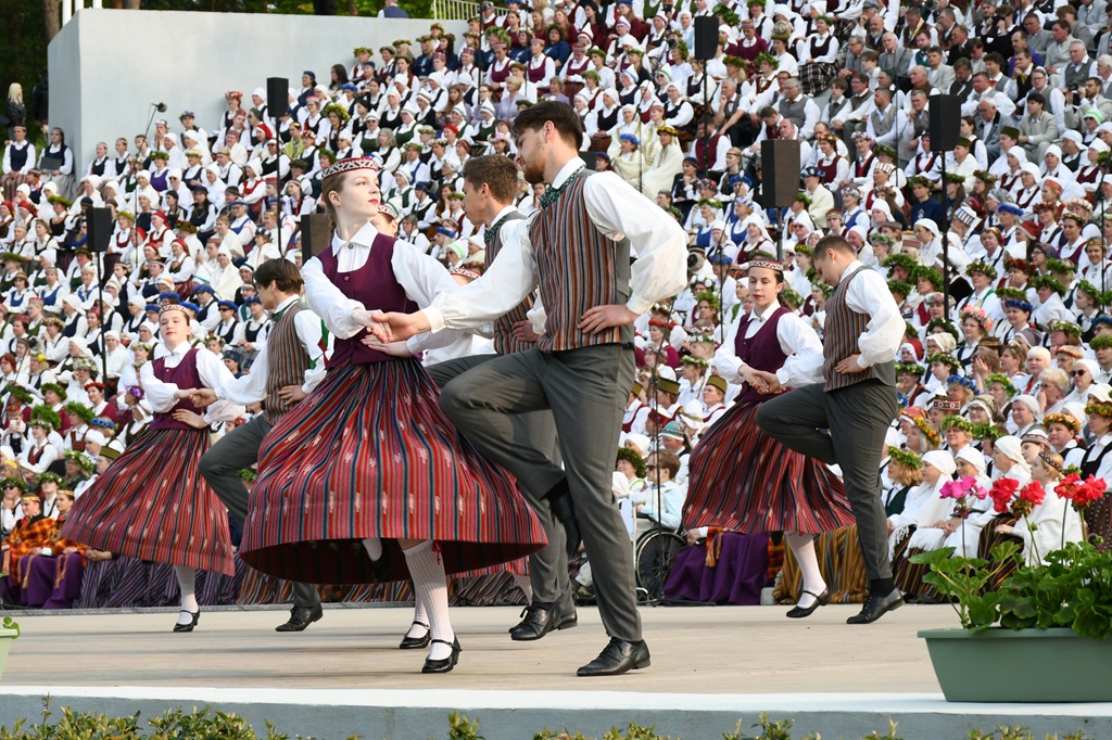 Tradicionālie dejotāji košās svītrainās svārkos un vestēs enerģiski uzstājas uz skatuves, ko ieskauj liela auditorija krāsainos tērpos.