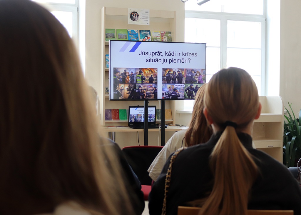 Cilvēku grupa, kas skatās uz lielu ekrānu, kurā redzami attēli un teksts latviešu valodā. Telpā ir gaiši logi un grāmatu plaukti, kas rada mācību atmosfēru.