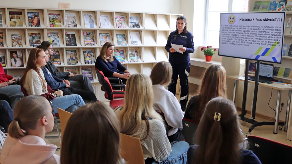 Sieviete formas tērpā stāv sēdošas grupas priekšā un ekrānā pasniedz informāciju. Fonā plaukti ar žurnāliem. Atmosfēra ir uzmanīga un izglītojoša.