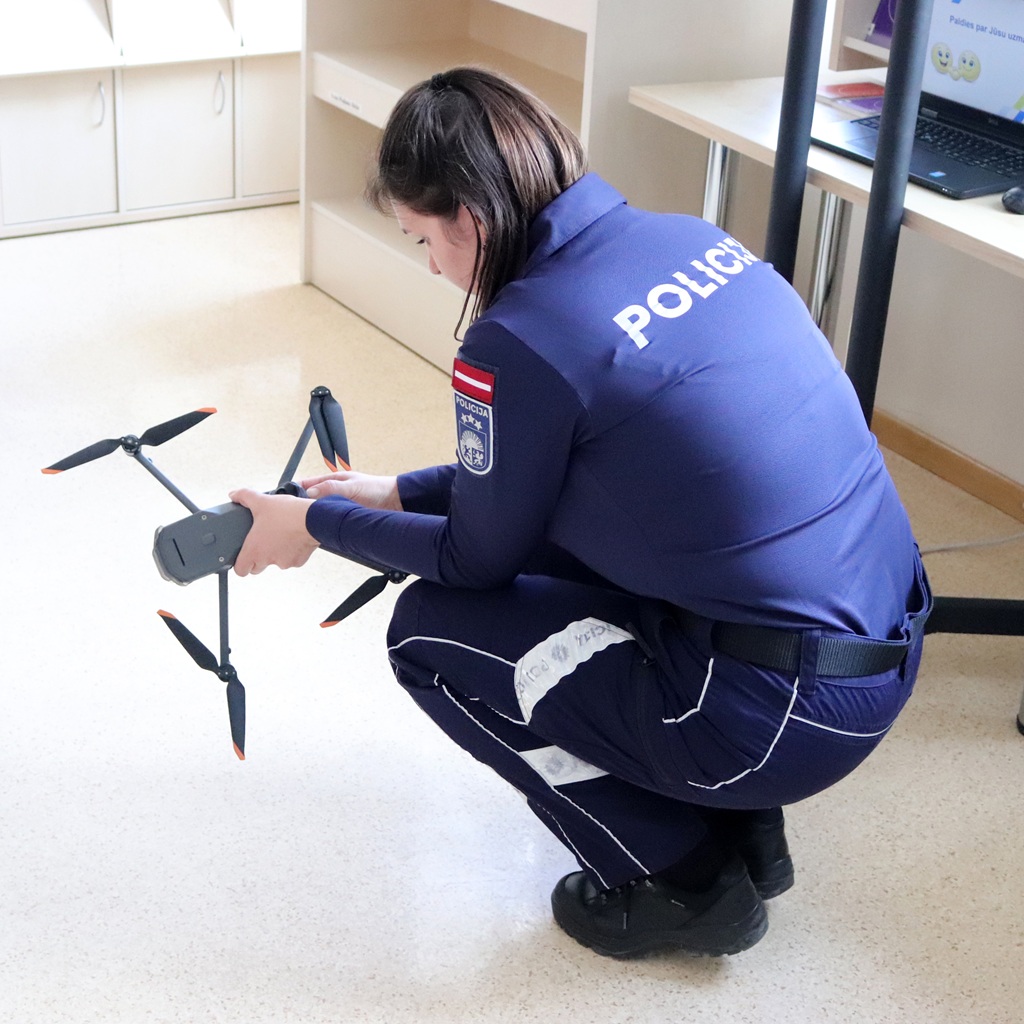 Policijas darbinieks formas tērpā tupē telpās, regulējot drona propellerus. Vide ir gaiša istaba ar plauktiem un galdu, kas liecina par tehnoloģiju uzstādīšanu.