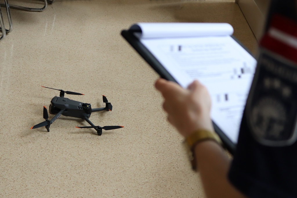 Cilvēks, stāvot blakus nelielam melnam dronam uz bēšas grīdas, pēta planšeti ar dokumentiem. Apkārtne atgādina apmācību vai novērtēšanas sesiju.