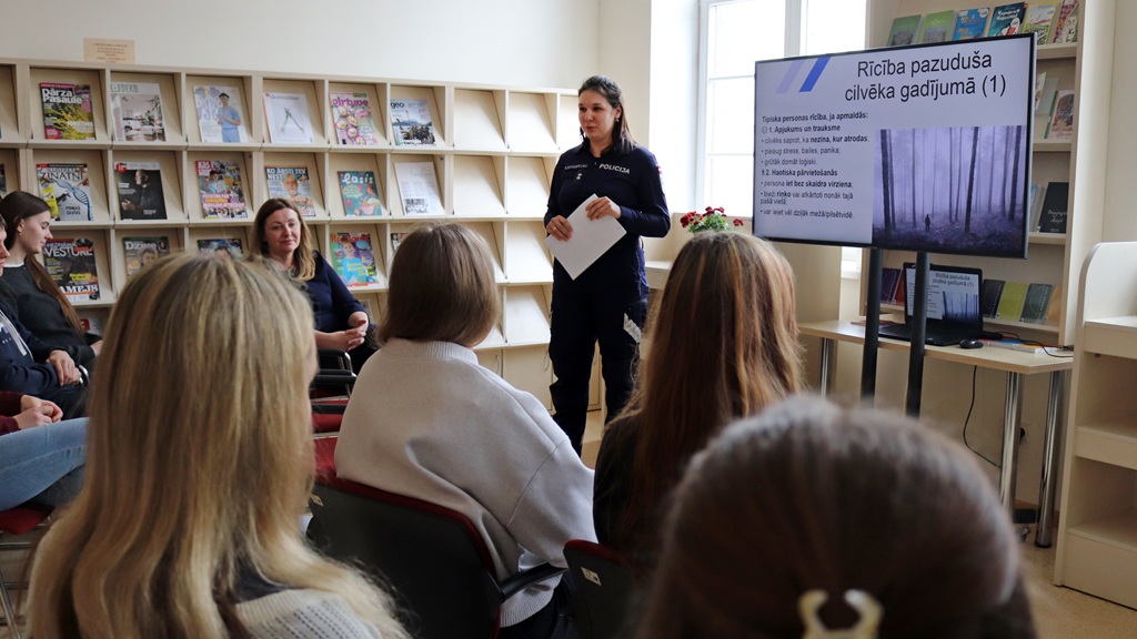 Sieviete policijas formas tērpā stāv grupas priekšā, kas sēž telpā ar grāmatu plauktiem, un sniedz prezentāciju. Ekrānā informācija tiek rādīta latviešu valodā.