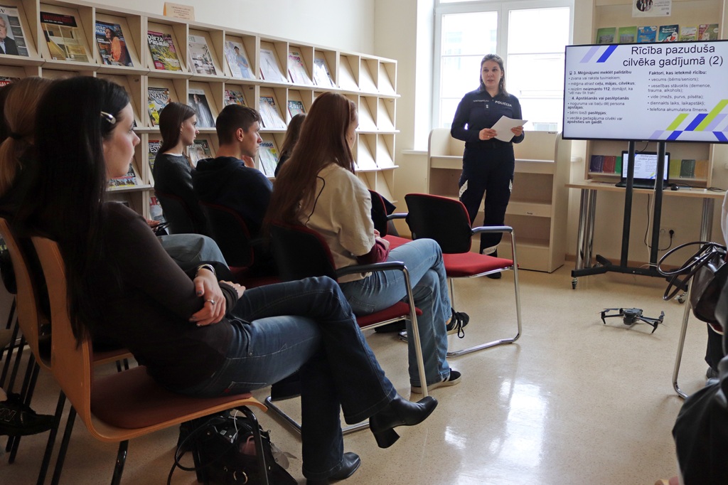 Sieviete uzstājas ar uzmanīgas auditorijas priekšā bibliotēkā, kuras plauktos ir pilni žurnāli. Uz liela ekrāna tiek rādīts teksts latviešu valodā. Atmosfēra ir koncentrēta un izglītojoša.