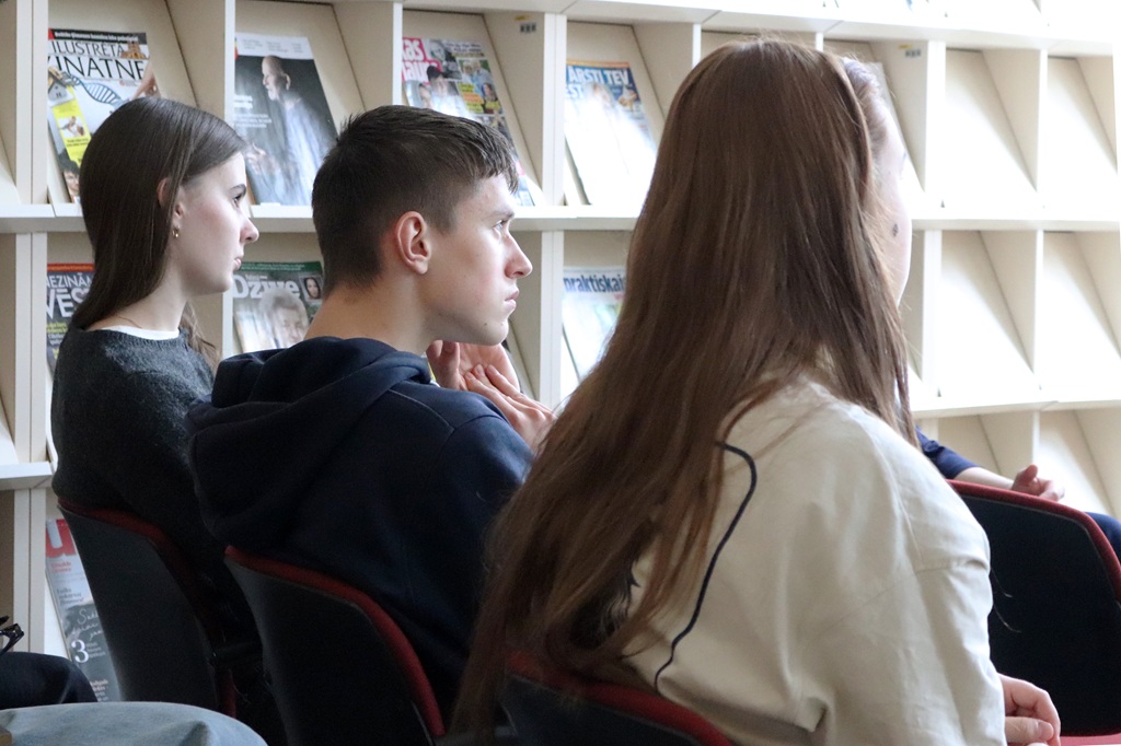 Jauniešu grupa uzmanīgi sēž bibliotēkā, fonā plauktos izlikti žurnāli. Valda koncentrēta un iesaistīta atmosfēra.