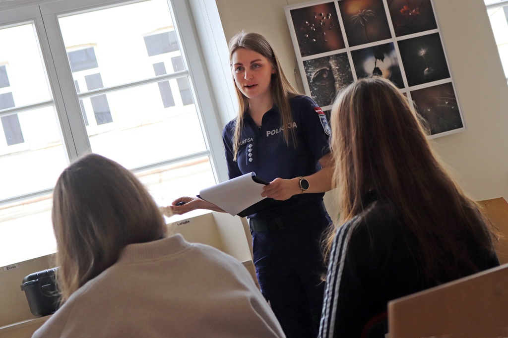 Policists formas tērpā gaišā telpā diviem sēdošiem cilvēkiem sniedz prezentāciju. Sienu rotā foto kolāža; atmosfēra ir izglītojoša.