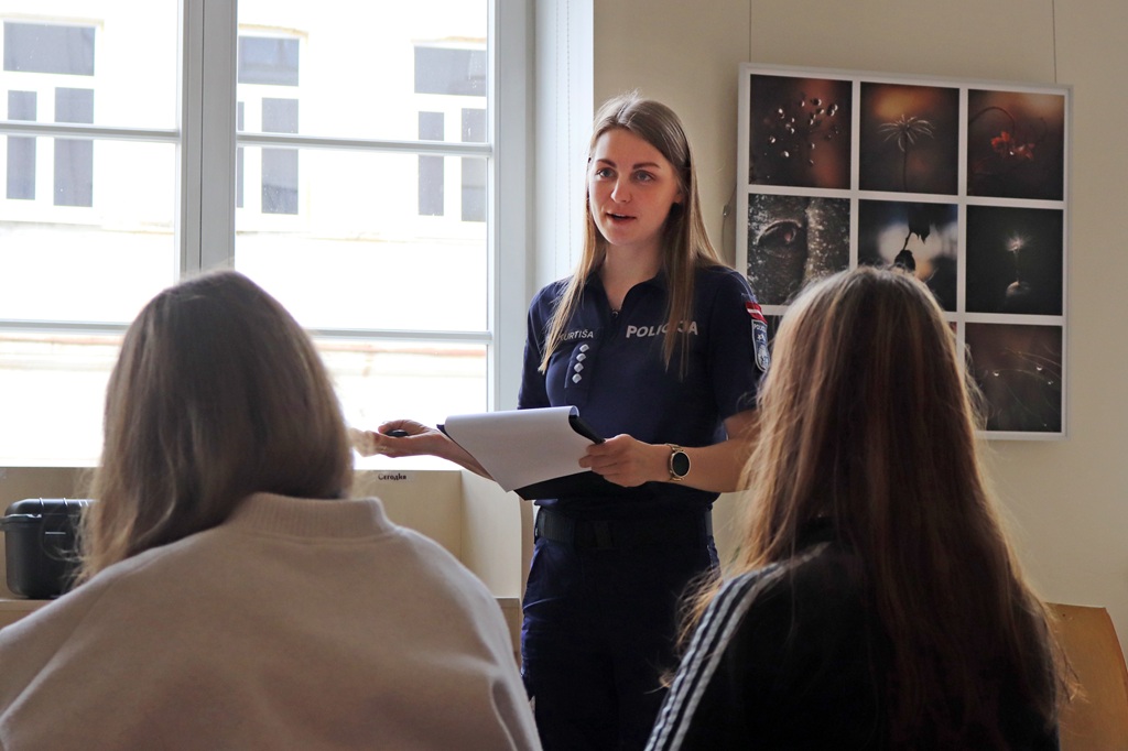 Policists formas tērpā gaišā telpā diviem sēdošiem cilvēkiem sniedz prezentāciju. Sienu rotā foto kolāža; atmosfēra ir izglītojoša.