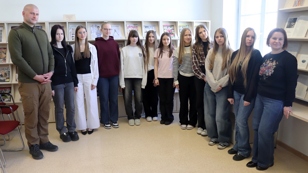 Cilvēku grupa stāv telpās, smaidot, uz fona, kurā ir grāmatu plaukti, kas pilni ar žurnāliem. Atmosfēra ir neformāla un draudzīga.