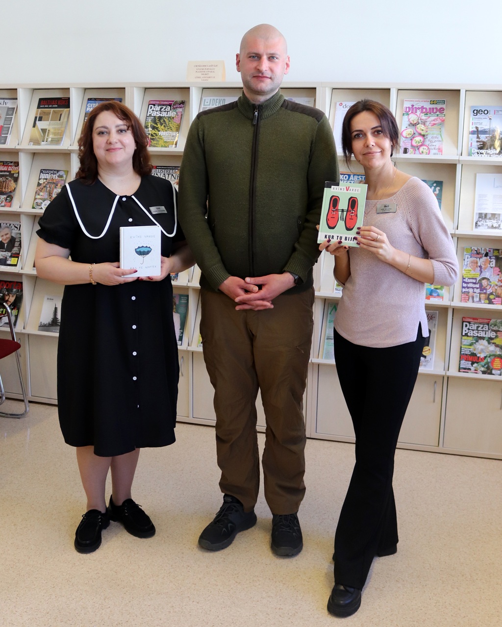 Trīs cilvēki smaidoši stāv bibliotēkā. Sieviete kreisajā pusē tur grāmatu baltos vākos, bet sieviete labajā pusē tur grāmatu ar sarkanām kurpēm uz vāka. Starp viņiem stāv vīrietis. Fonā redzami plaukti, kas pilni ar žurnāliem.