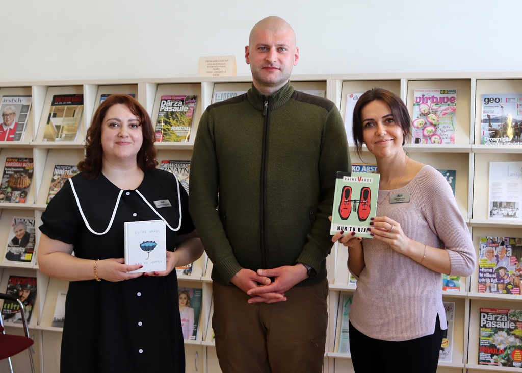 Trīs cilvēki stāv bibliotēkā ar grāmatām rokās. Fonā plauktos izvietoti žurnāli.