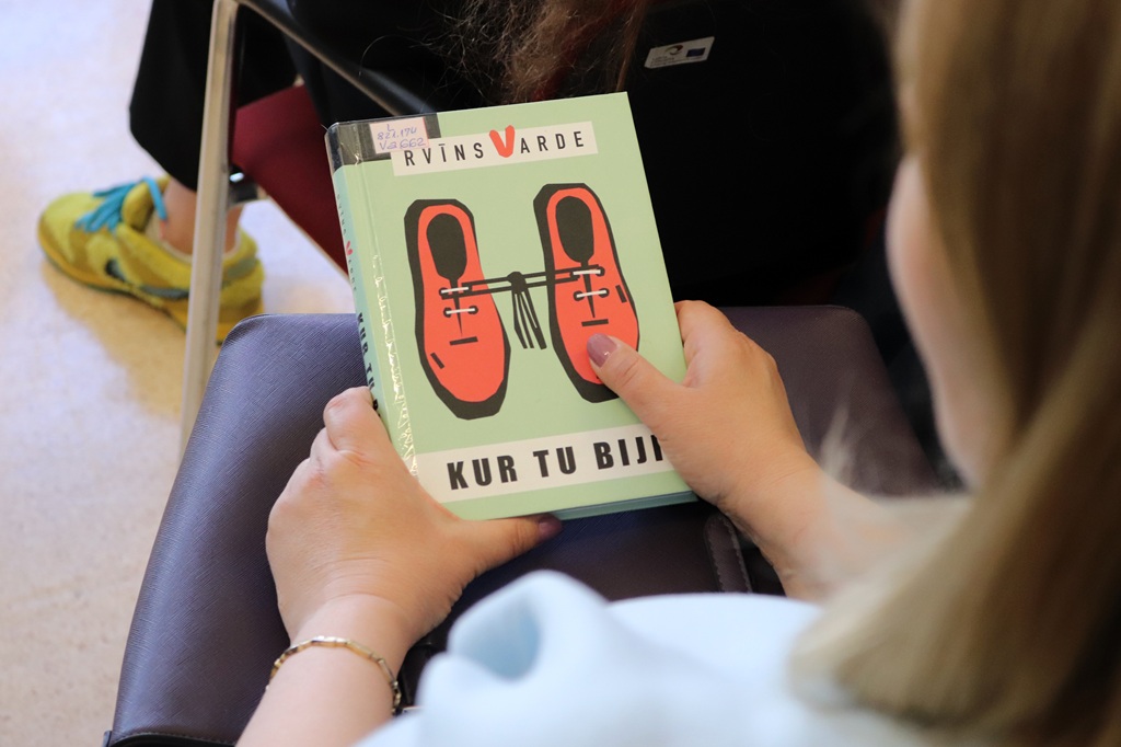 Cilvēks tur rokās grāmatu ar nosaukumu "Kur Tu Biji", uz kuras vāka attēlotas kopā sasietas sarkanas kurpes. Netālu sēž vēl viens cilvēks dzeltenās kurpēs, radot mājīgu atmosfēru.