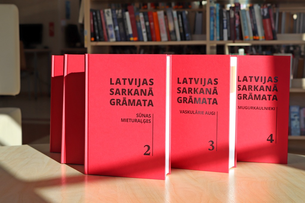 Uz saules apspīdēta galda ir izliktas sarkanas grāmatas ar nosaukumu "Latvijas Sarkanā Grāmata". Grāmatas ir numurētas ar 2, 3 un 4, fonā — grāmatu plaukts.
