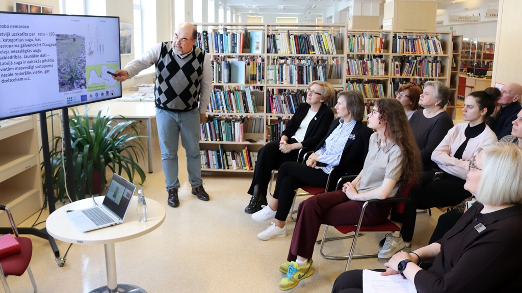 Vīrietis rūtainā džemperī bibliotēkā lielā ekrānā rāda informāciju sēdošai cilvēku grupai. Fonā redzami grāmatu plaukti. Tonis ir izglītojošs.