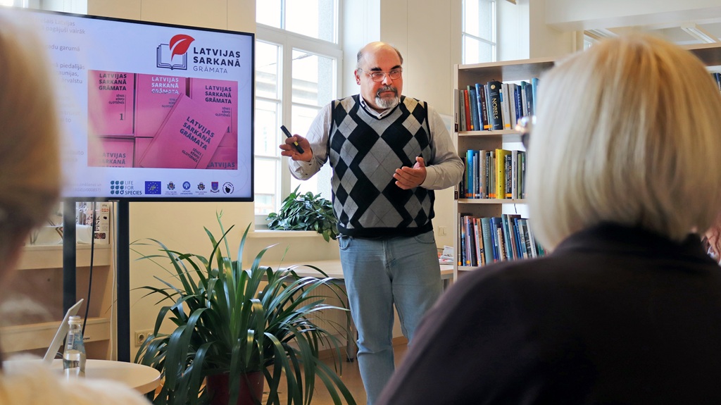 Vīrietis bibliotēkā sniedz prezentāciju, žestikulējot ar roku. Viņam blakus ekrānā redzama "Latvijas Sarkanā Grāmata". Fonā grāmatu plaukti un augi.