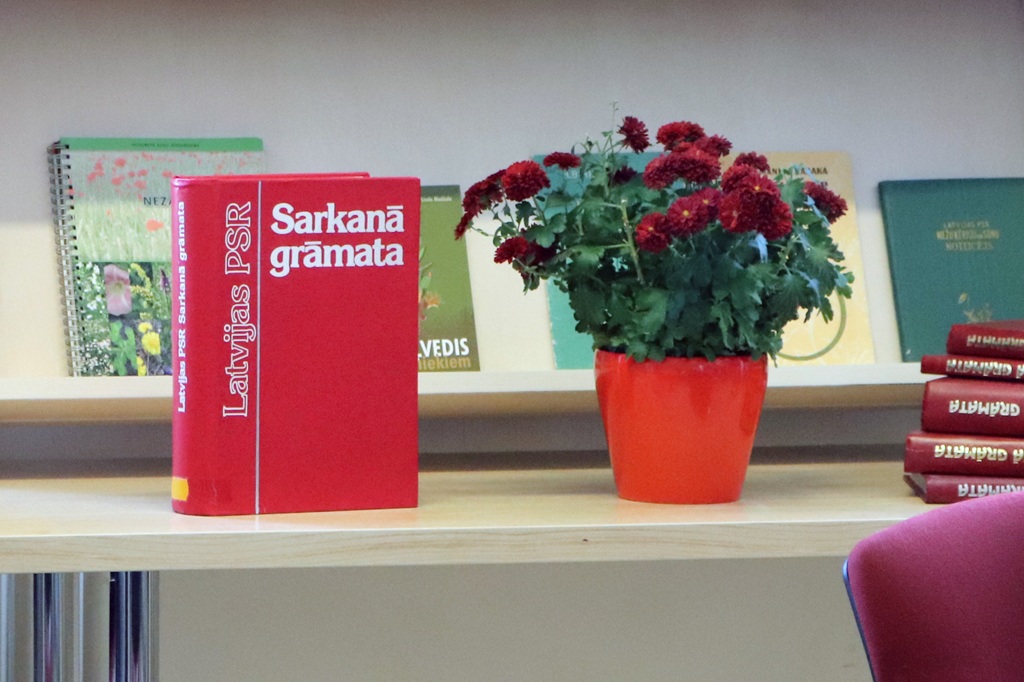 Plauktā blakus podam ar bordo ziediem atrodas liela sarkana grāmata ar nosaukumu "Sarkanā grāmata". Fonā redzamas vairākas zaļas un brūnas grāmatas.