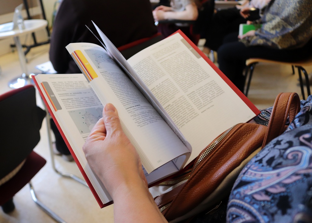 Cilvēks pāršķirsta biezu, cietos vākos iesietu grāmatu. Aina pauž fokusu un iesaisti, fonā redzamas izplūdušas figūras.