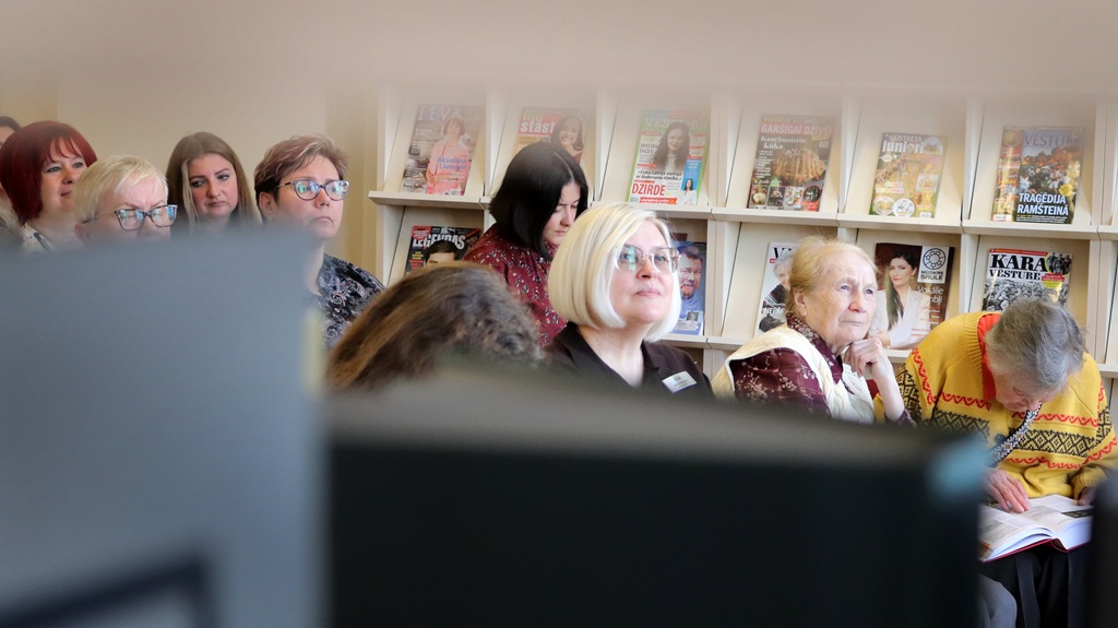 Bibliotēkā uzmanīgi sēž cilvēku grupa, aiz viņiem plauktos izlikti žurnāli. Atmosfēra ir koncentrēta un iesaistīta.