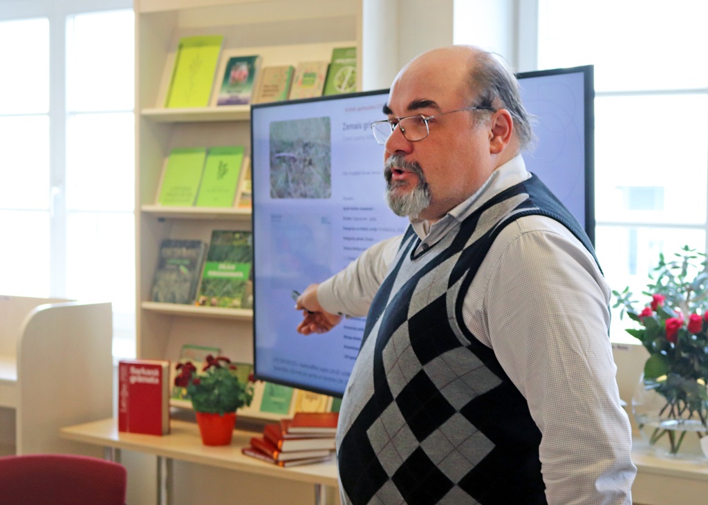 Vīrietis džempera vestē norāda uz lielu ekrānu, kurā redzams teksts un attēli, ko ieskauj grāmatu plaukti un augi, un sniedz izglītojošu prezentāciju.