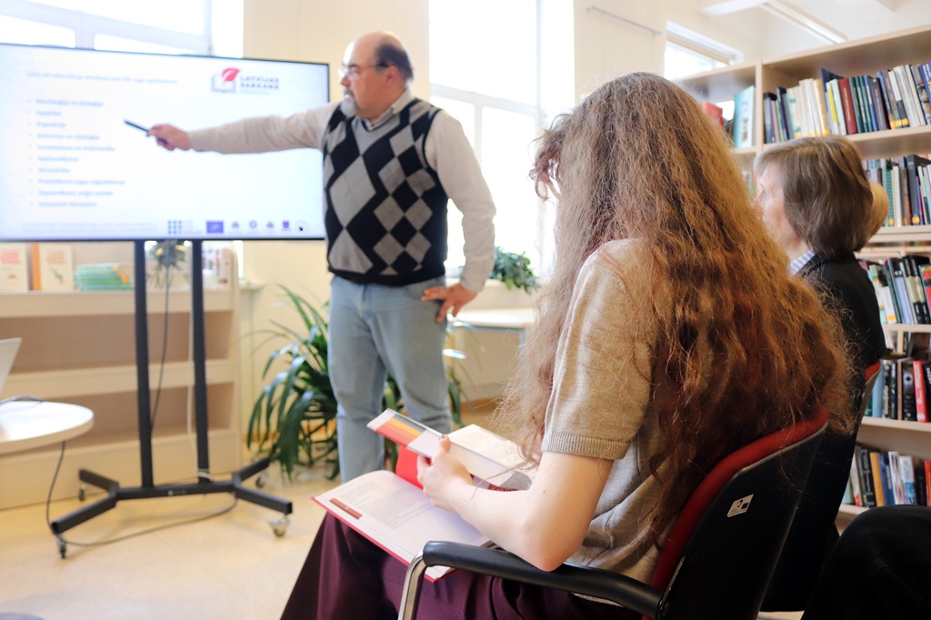 Vīrietis džempera vestē norāda uz prezentāciju ekrānā bibliotēkā. Divas sievietes sēž viņam pretī un ar koncentrētām sejas izteiksmēm pieraksta piezīmes.