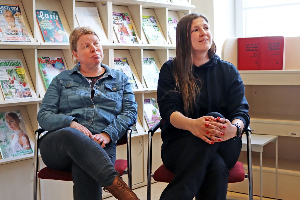 Divas sievietes sēž krēslos bibliotēkā. Viena ģērbusies džinsa jakā, otra – melnā kapučjakā. Aiz viņām plauktos sarindotas grāmatas un žurnāli.