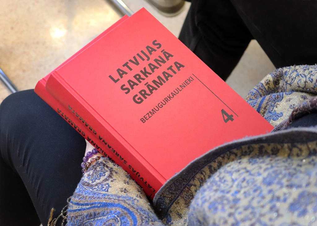 Cilvēkam rokās ir spilgti sarkana grāmata ar nosaukumu "Latvijas Sarkanā Grāmata: Bezmugurkaulnieki 4." Vāka treknais teksts kontrastē ar cilvēka rakstaino zilo un zelta audumu.