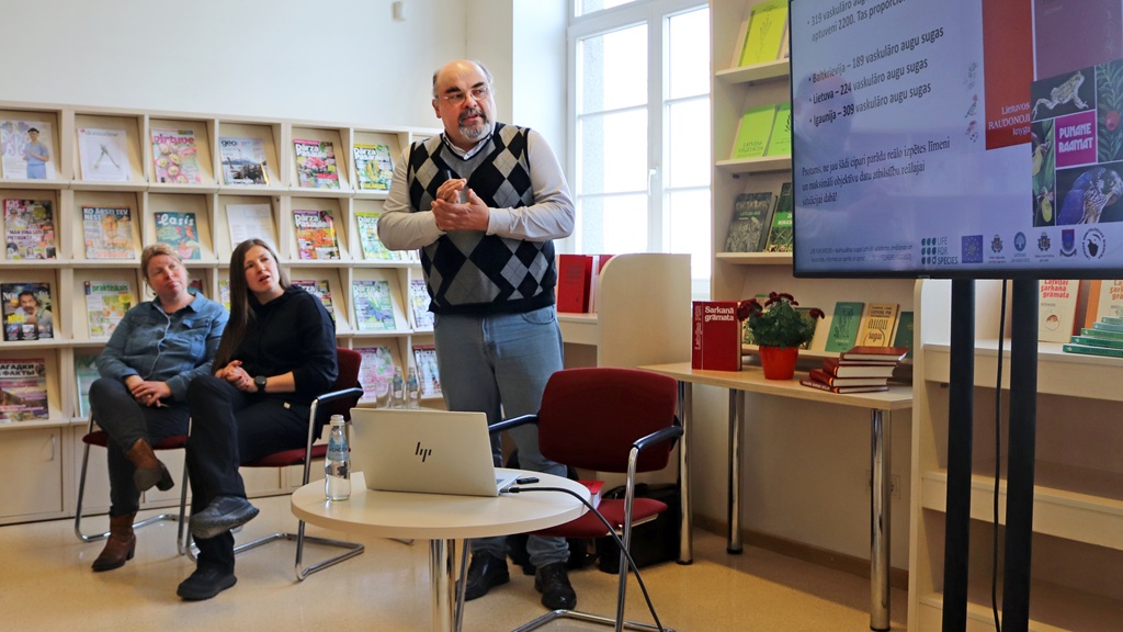 Vīrietis džempera vestē uzstājas bibliotēkā, žestikulējot uz ekrānu, kurā redzami slaidi. Fonā uzmanīgi sēž divi cilvēki. Vide ir mierīga un koncentrēta.