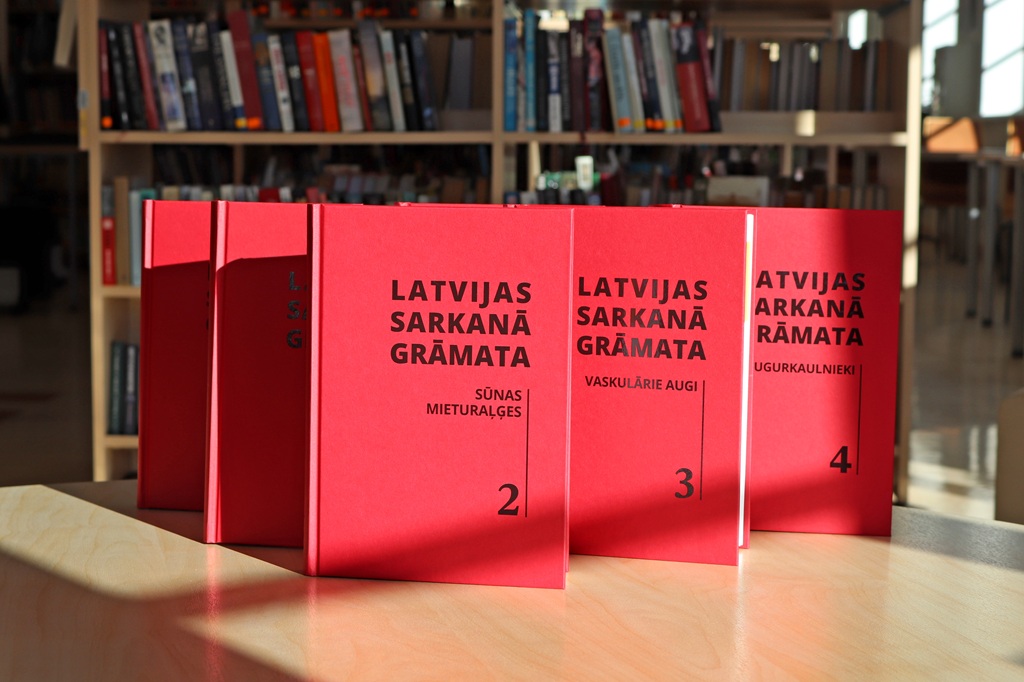 Bibliotēkā uz koka galda sarindotas sarkanas grāmatas ar nosaukumu "Latvijas Sarkanā Grāmata". Saules gaisma apspīd grāmatas, piešķirot tām akadēmisku noskaņu.
