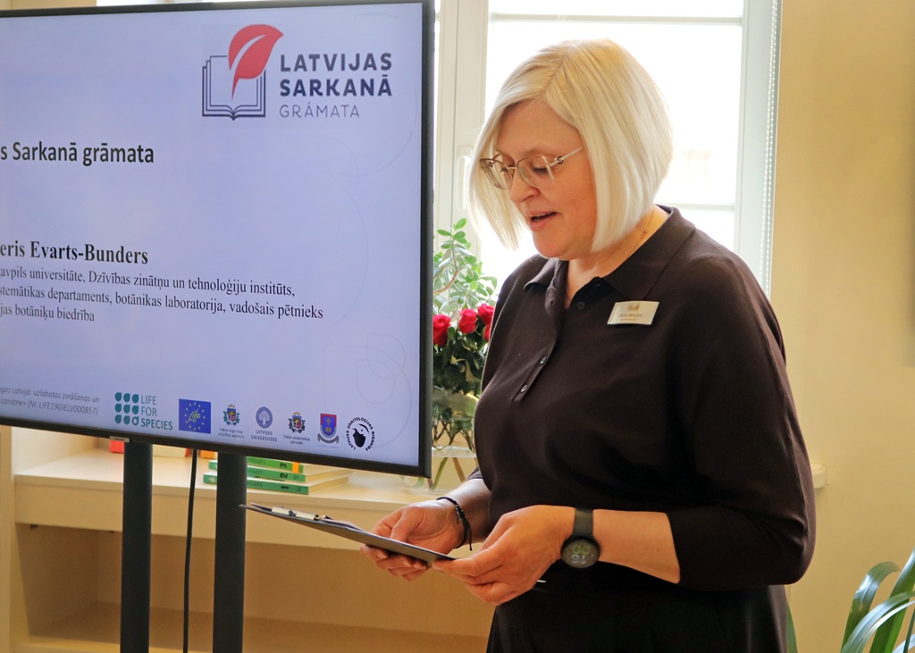 Sieviete ar īsiem blondiem matiem un brillēm lasa no planšetes gaišā istabā. Aiz viņas ir ekrāns ar uzrakstu "Latvijas Sarkanā Grāmata".