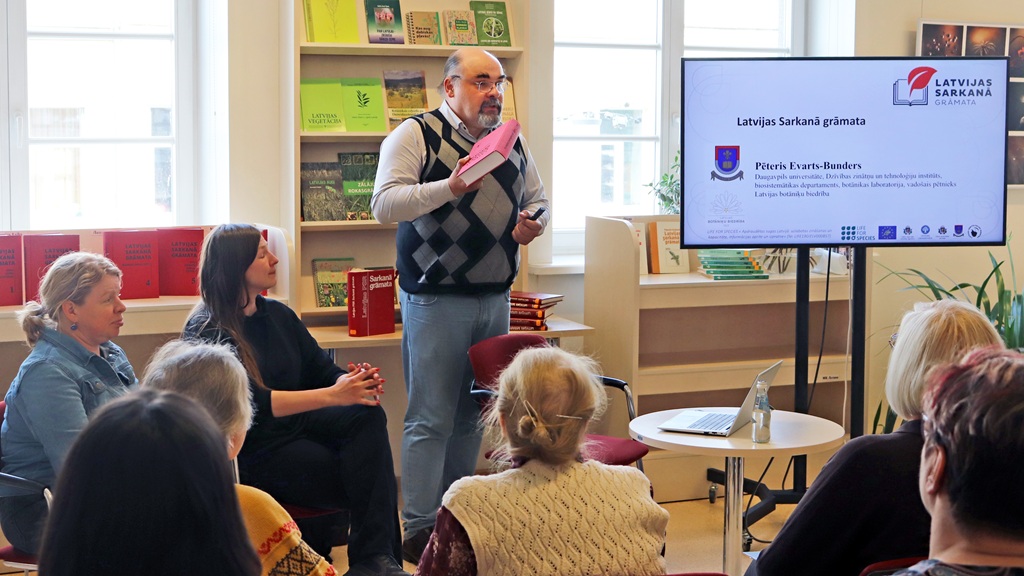 Runātājs bibliotēkā tur rokās rozā grāmatu, uzrunājot uzmanīgu auditoriju. Uz ekrāna redzama prezentācija "Latvijas Sarkanā grāmata". Noskaņojums ir izglītojošs.