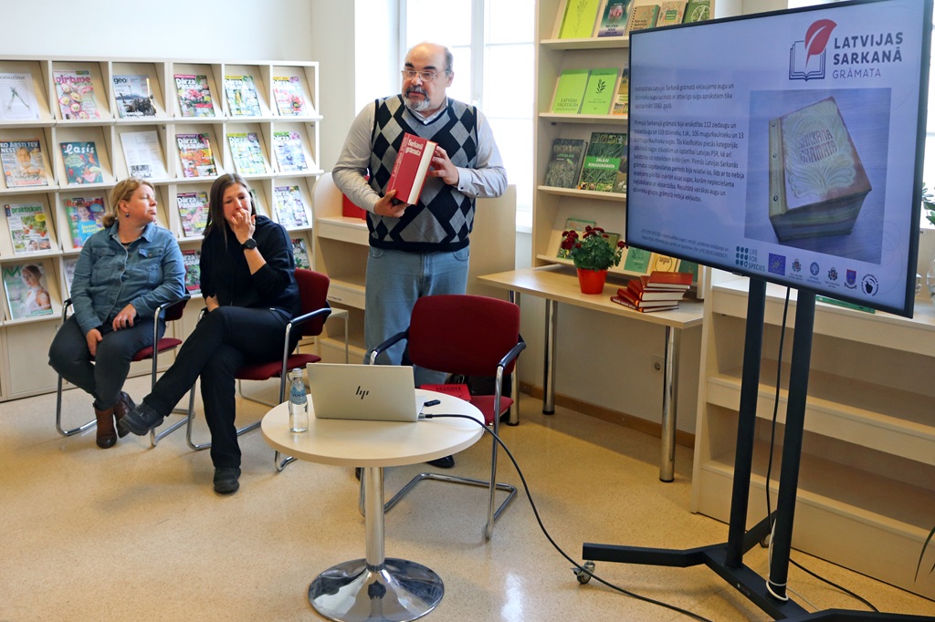 Vīrietis bibliotēkā tur rokās sarkanu grāmatu. Divas sievietes uzmanīgi sēž. Ekrānā redzama informācija par "Latvijas Sarkano Grāmatu". Fonā plaukti ar žurnāliem.