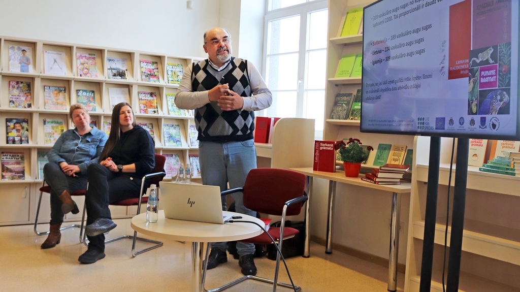 Vīrietis bibliotēkā uzstājas ar projektoru; divi auditorijas locekļi uzmanīgi klausās. Telpa ir gaiša, gar sienām rotā grāmatas un žurnāli.