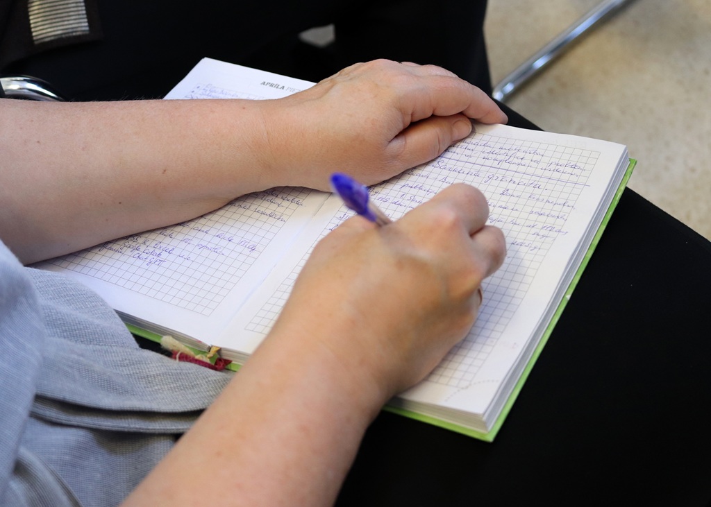 Tuvplāns ar rokām, kas raksta piezīmju grāmatiņā ar rūtiņu papīru, izmantojot violetu pildspalvu. Vide izskatās relaksēta un koncentrēta, kas liecina par piezīmju veikšanu vai dienasgrāmatas ierakstīšanu.