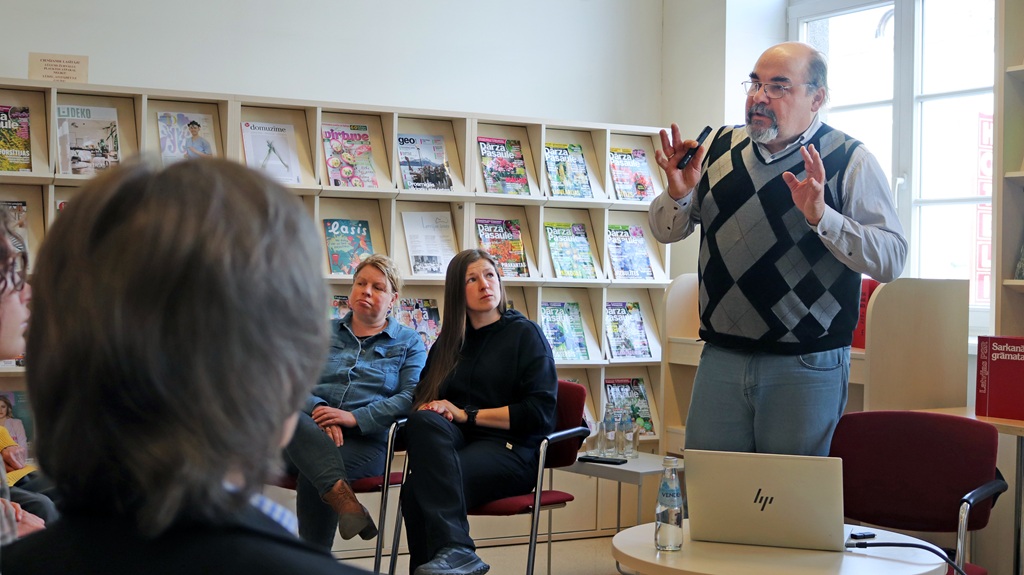 Vīrietis žestikulē, uzrunājot nelielu auditoriju bibliotēkā. Klātesošie uzmanīgi sēž žurnālu plauktu priekšā, radot aizraujošu, izglītojošu atmosfēru.