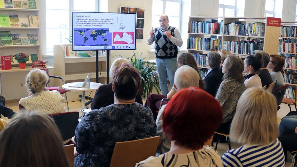 Vīrietis bibliotēkā uzmanīgai auditorijai sniedz prezentāciju. Viņš norāda uz lielu ekrānu, kurā redzama karte un dati, ko ieskauj grāmatu plaukti.