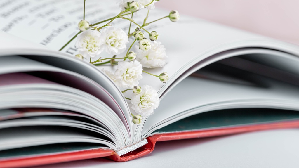 Uz līdzenas virsmas guļ atvērta grāmata ar sarkanu vāku, un uz tās lapām atpūšas mazi balti ziediņi.