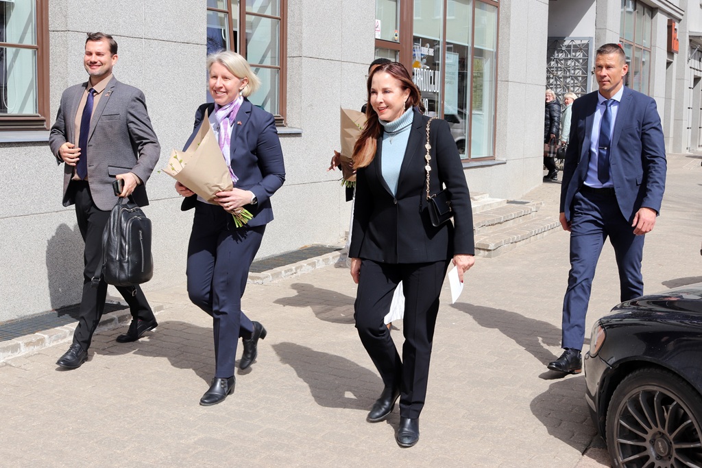 Saulainā dienā ārā pastaigājas četru profesionāli ģērbtu cilvēku grupa, divi tur pušķus, izskatoties dzīvespriecīgi un iesaistīti.