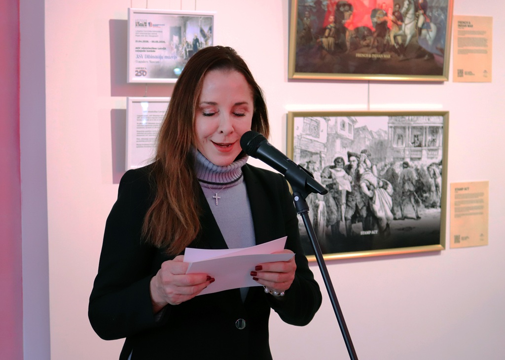 Sieviete melnā žaketē lasa no papīra pie mikrofona. Viņa stāv blakus ierāmētām gleznām, radot siltu un saistošu atmosfēru.