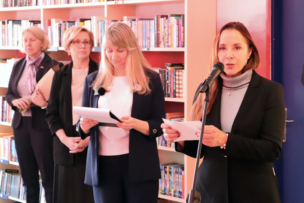 Četras sievietes stāv bibliotēkā pie grāmatu plauktiem. Viena sieviete runā mikrofonā, bet pārējās tur papīrus, radot iespaidu par oficiālu pasākumu.