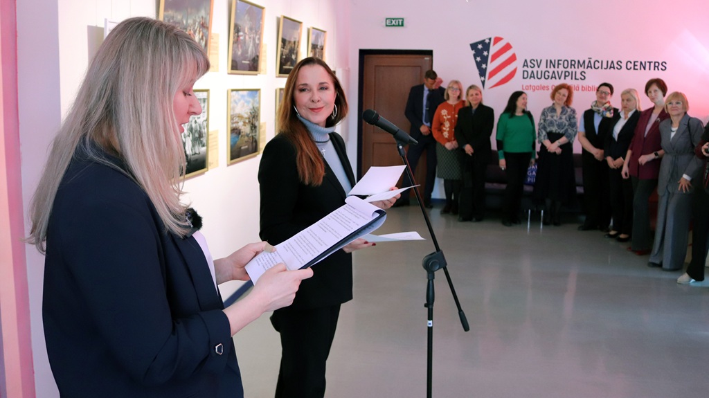 Sieviete runā pie mikrofona, turot rokās papīrus, spilgti apgaismotā telpā, aiz viņas ir sapulcējušies cilvēki. Uz plakāta ir uzraksts "ASV Informācijas Centrs Daugavpils".