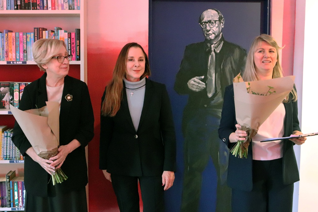 Trīs sievietes stāv istabā, divas tur pušķus, fonā – grāmatu plaukti un liela vīrieša glezna, radot formālu un svinīgu noskaņu.