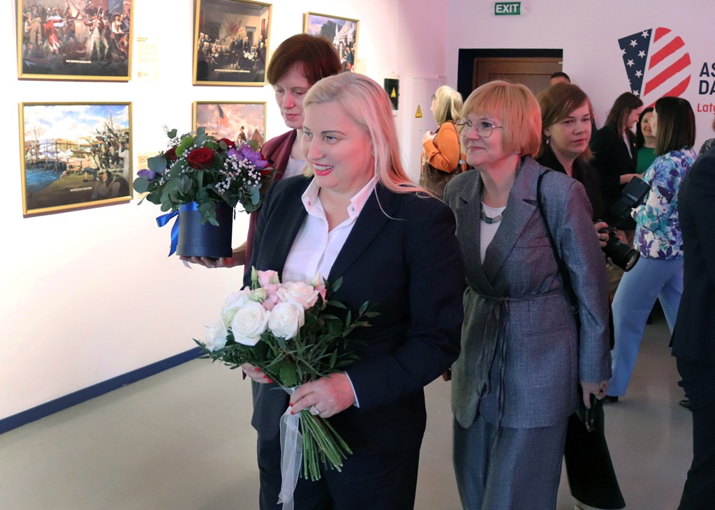 Sieviešu grupa pastaigājas pa mākslas galeriju, viena no tām stāv priekšā un tur rokās baltu rožu pušķi. Viņas izskatās laimīgas un ir ģērbušās svinīgi.